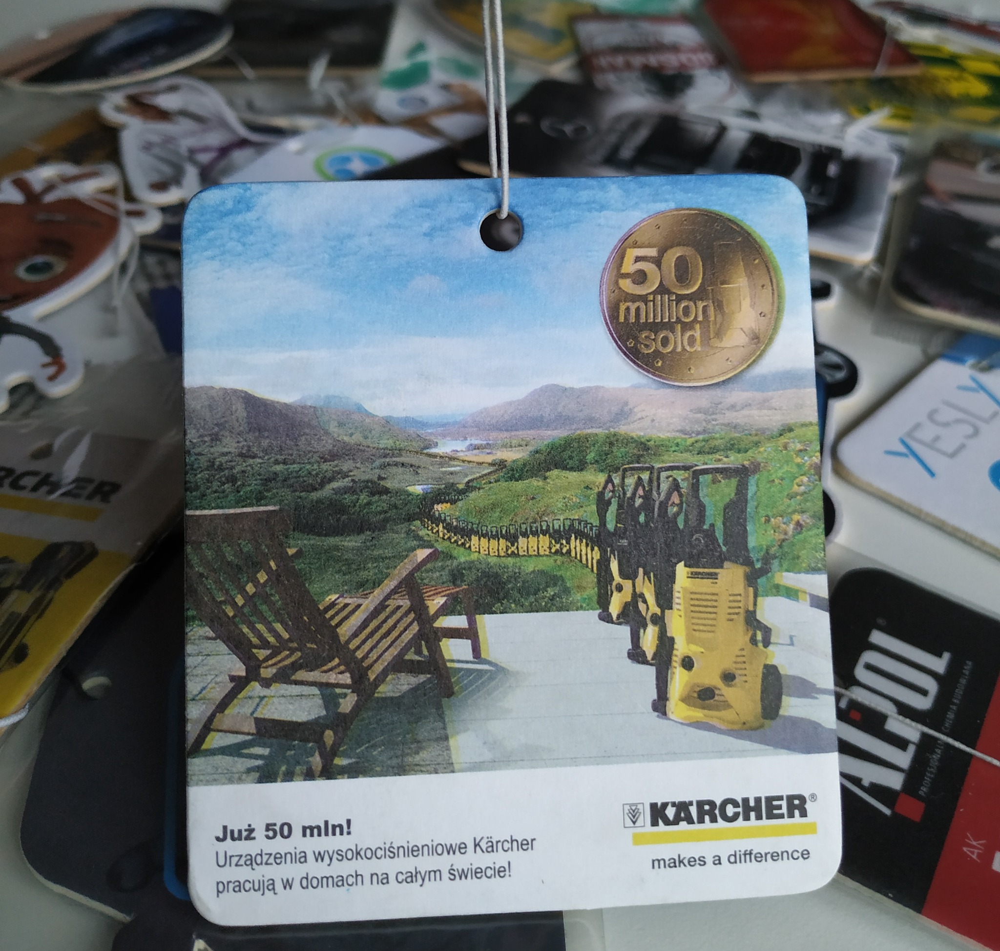 Zawieszka reklamowa Karcher z krajobrazem górskim, krzesłami ogrodowymi i rzędem myjek ciśnieniowych. W tle inne zawieszki reklamowe.