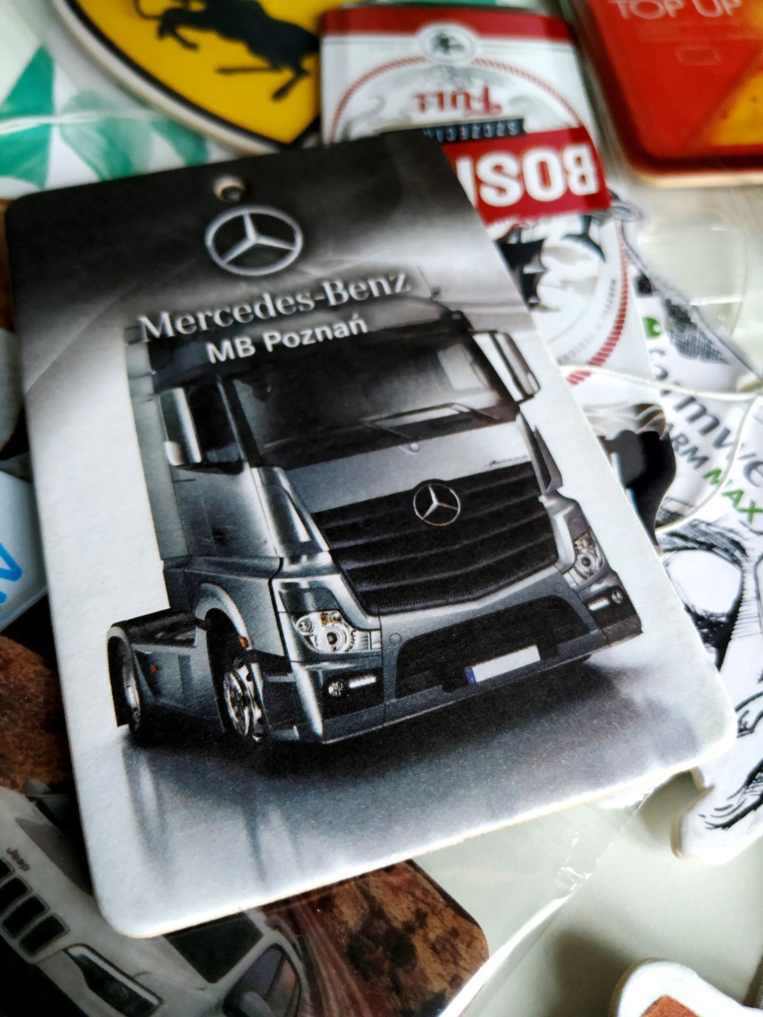 Brelok z grafiką srebrnego samochodu ciężarowego Mercedes-Benz MB Poznań, leżący na różnorodnym tle innych przedmiotów.