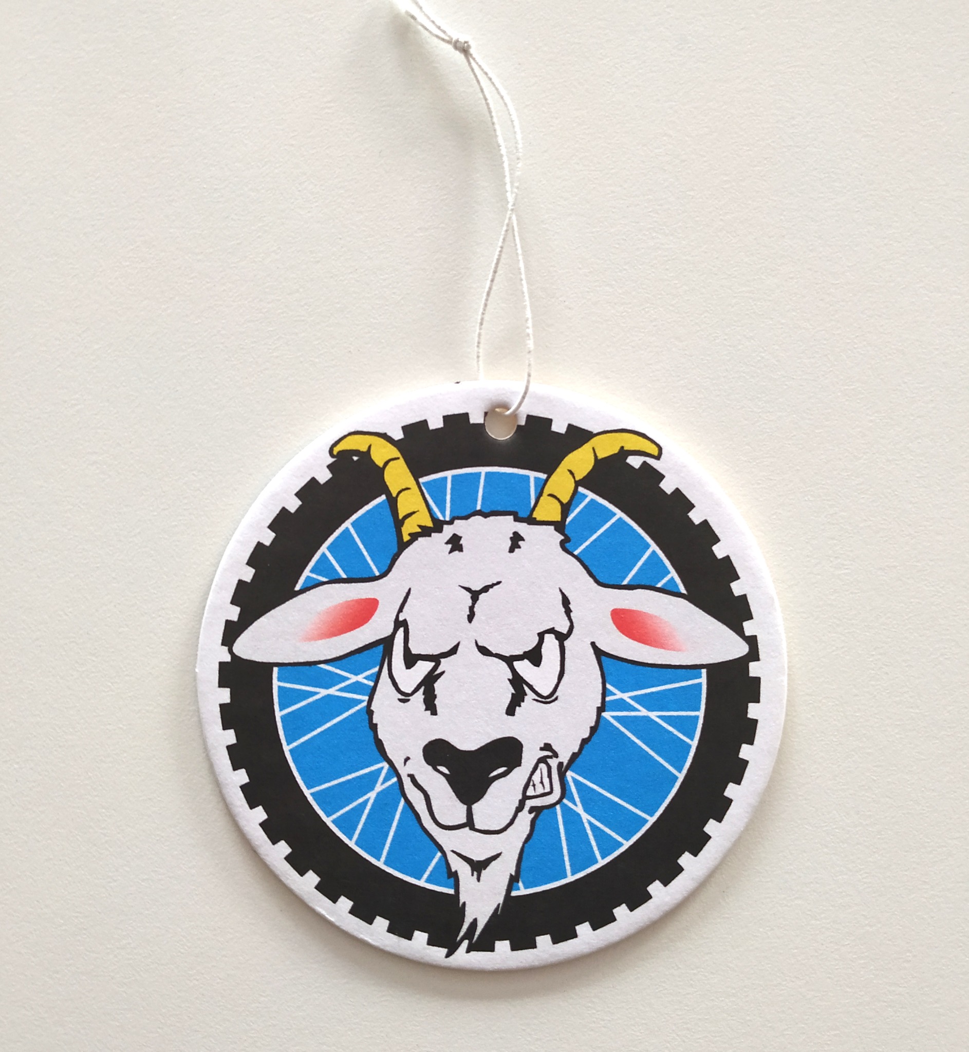 Okrągła zawieszka z ilustracją kozła o groźnym wyrazie twarzy na tle niebieskiego koła rowerowego z czarną oponą, zawieszona na białym sznurku.