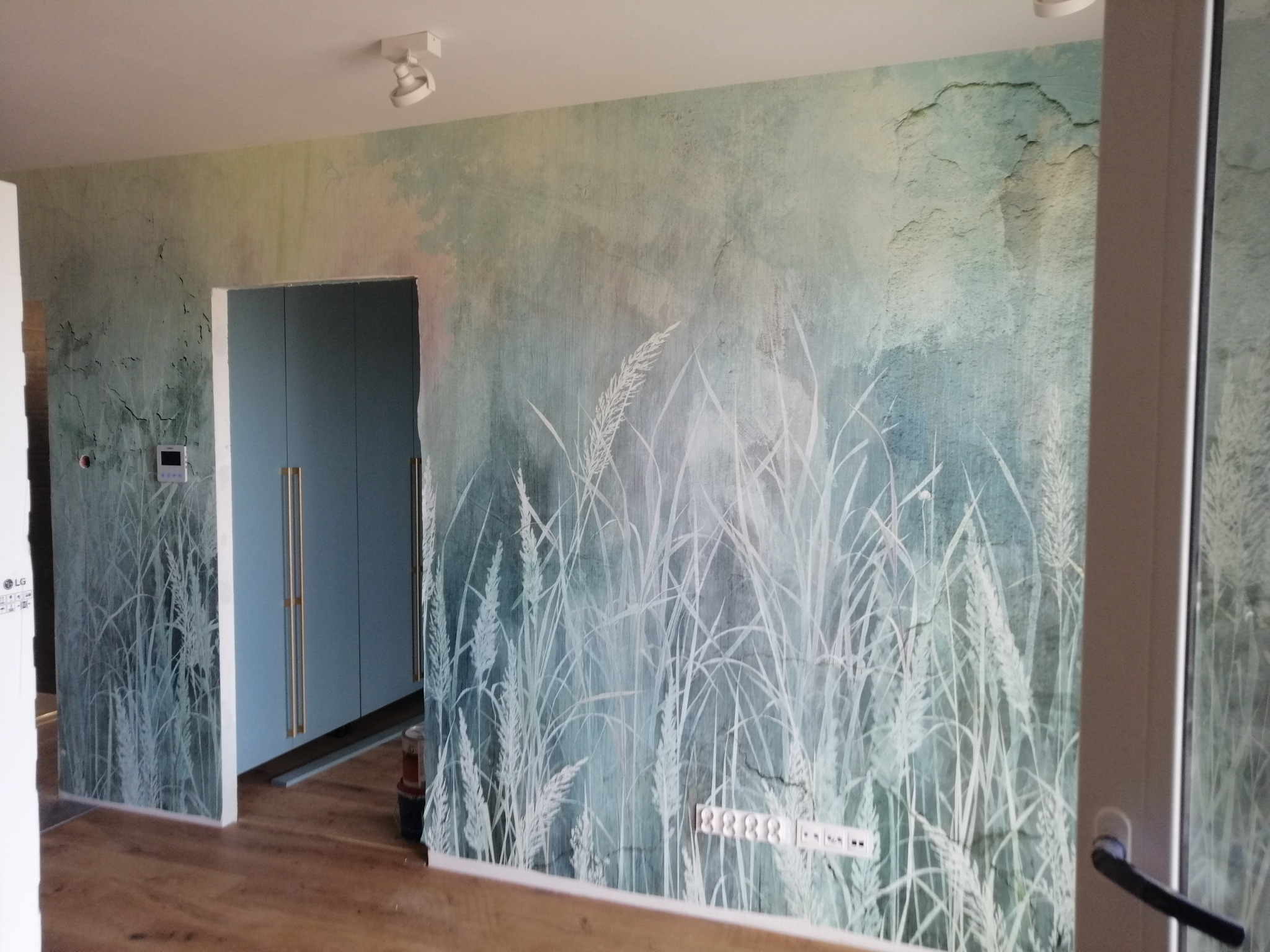 Pokój z tapetą w odcieniach zieleni i bieli, przedstawiającą motyw trawy, z widocznymi gniazdkami elektrycznymi i fragmentem szafy wnękowej w tle.