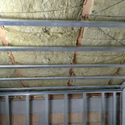 Montaż izolacji z wełny mineralnej pod sufitem z metalową konstrukcją, widoczne fragmenty ceglanej ściany z szarą zaprawą.