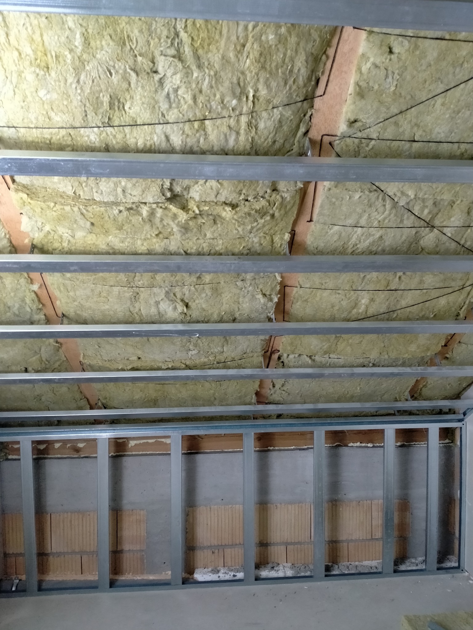 Montaż izolacji z wełny mineralnej pod sufitem z metalową konstrukcją, widoczne fragmenty ceglanej ściany z szarą zaprawą.