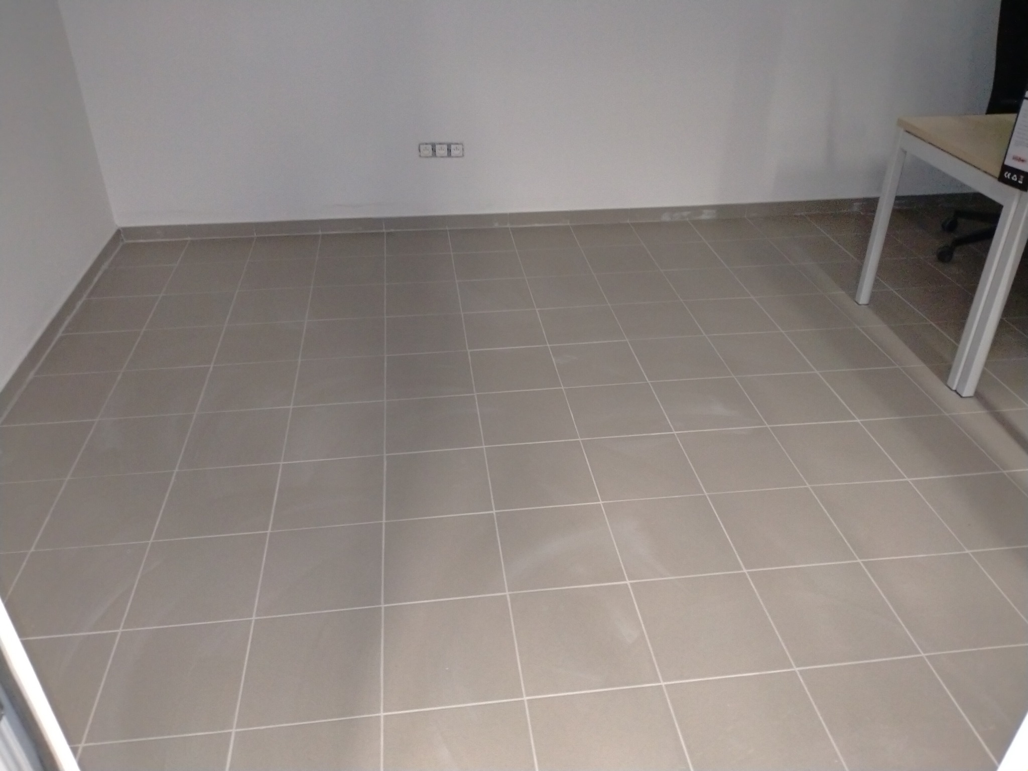 Ułożona, beżowa podłoga z płytek ceramicznych w pustym pomieszczeniu biurowym, widoczne gniazdka elektryczne na ścianie i fragment biurka z białymi nogami.
