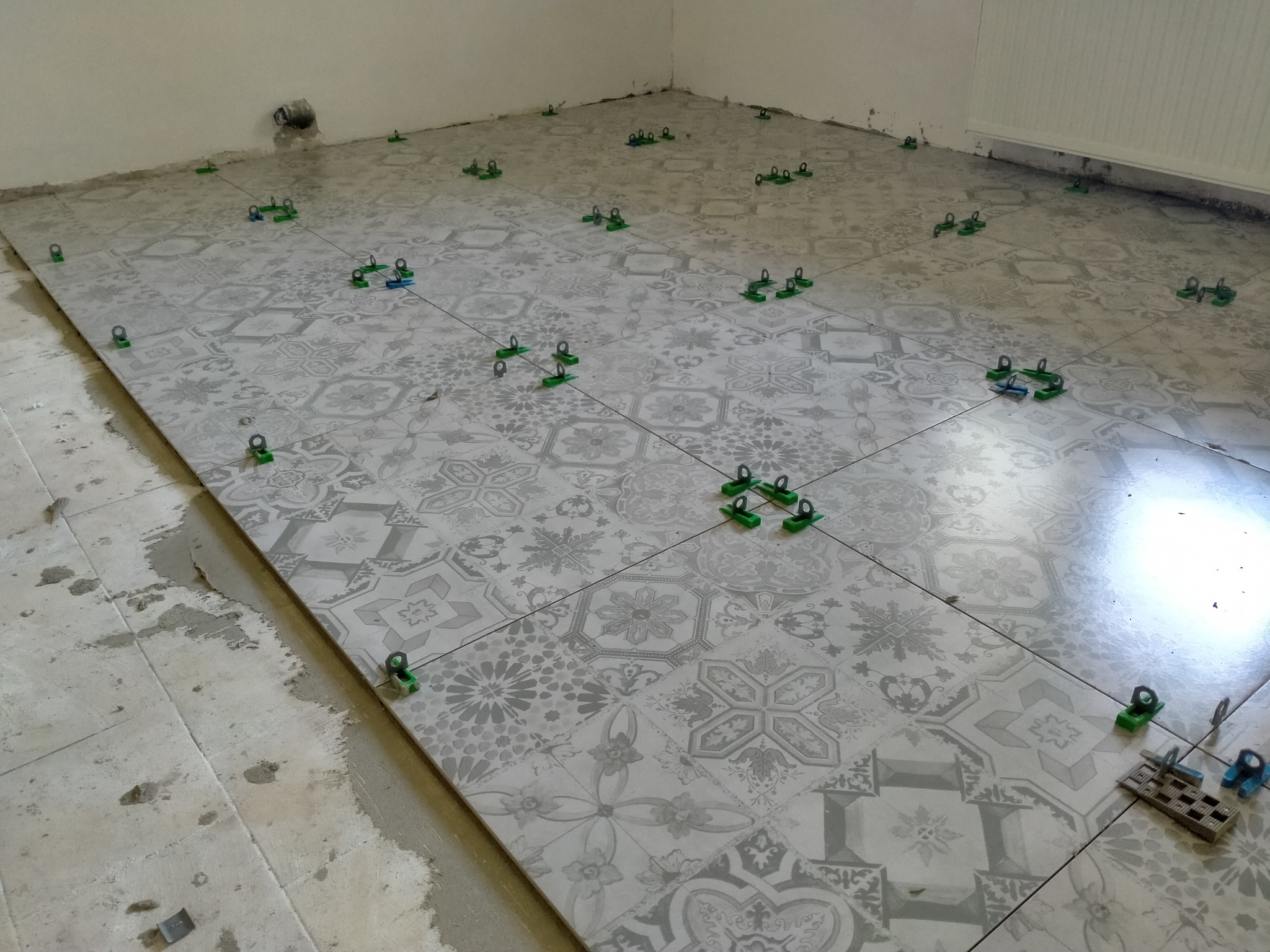 Układanie płytek podłogowych z dekoracyjnym wzorem, z użyciem zielonych klipsów poziomujących, widoczne niedociągnięcia przy ścianie i resztki kleju.