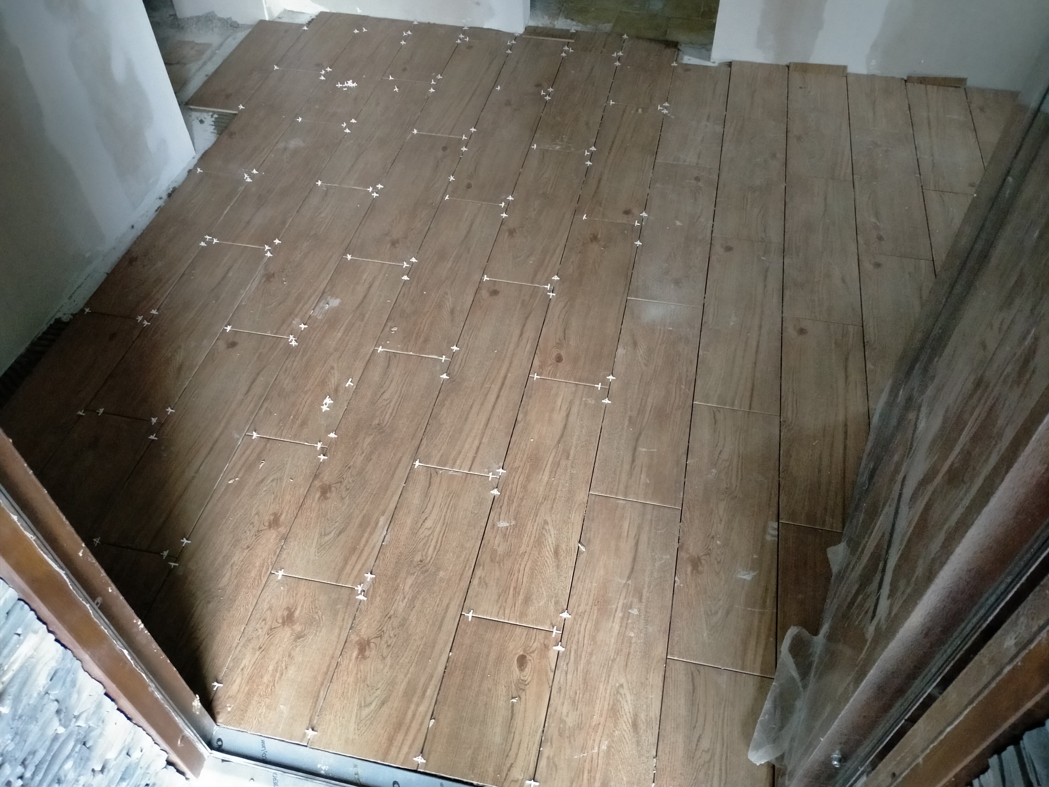 Układanie brązowych płytek podłogowych imitujących drewno z użyciem krzyżyków dystansowych, widok z góry na niedokończoną podłogę w trakcie remontu.