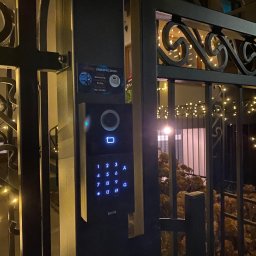 CONNECT ZABEZPIECZENIA - Nowoczesny wideodomofon Kenik z podświetlaną klawiaturą numeryczną, zamontowany na metalowej bramie wjazdowej, z widocznym logo firmy Connect Zabezpieczenia.