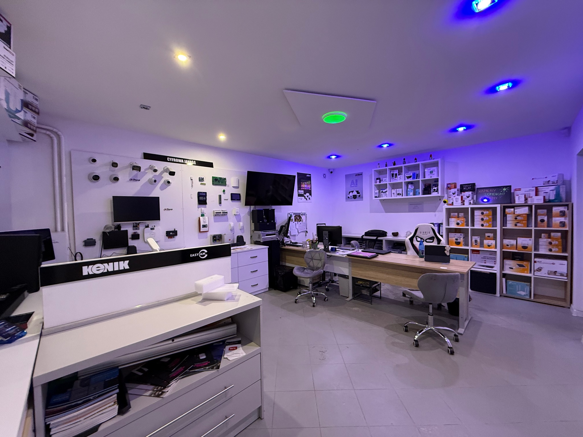 Salon z ekspozycją systemów zabezpieczeń: kamery, alarmy, domofony. Na ścianie monitory i urządzenia. Oświetlenie LED w odcieniach niebieskiego i zielonego.
