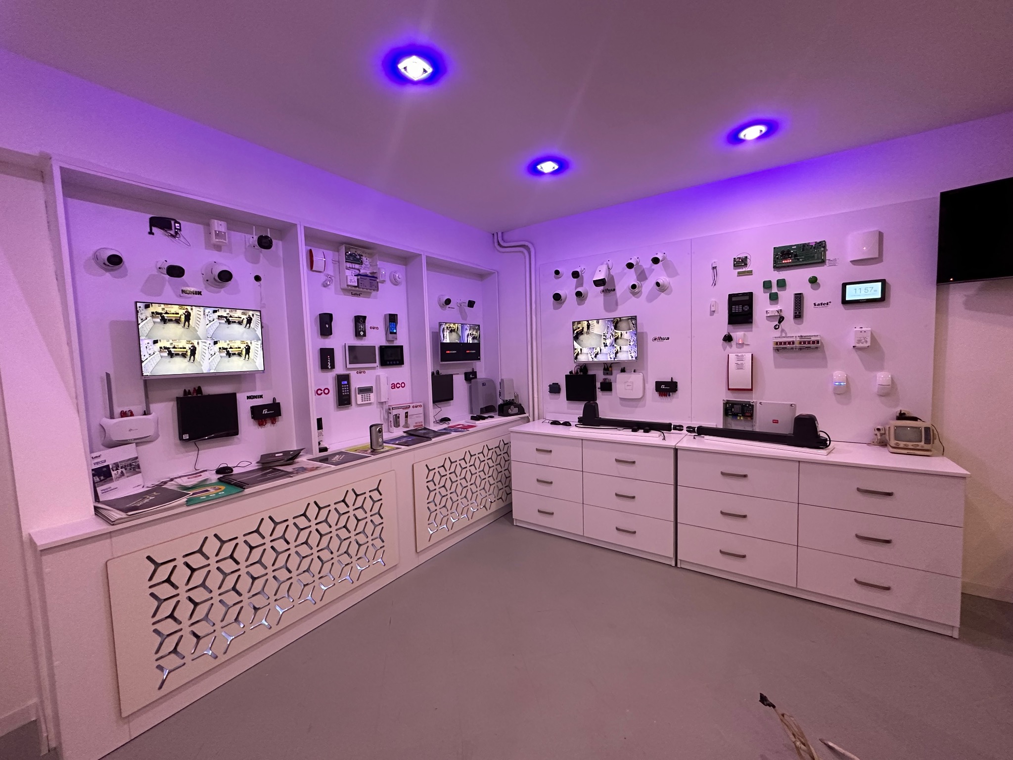 Salon ekspozycyjny z systemami zabezpieczeń: kamery, alarmy, domofony. Monitory prezentują obraz z kamer. Białe meble i ściany, fioletowe oświetlenie.
