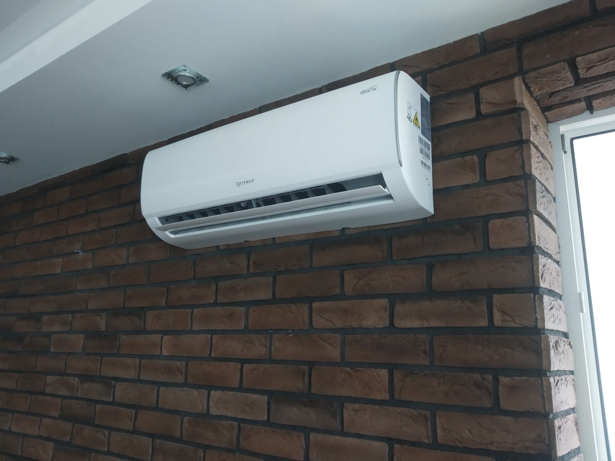 Klimatyzator marki Rotenso zamontowany na ścianie z cegły dekoracyjnej w pomieszczeniu z oświetleniem punktowym.