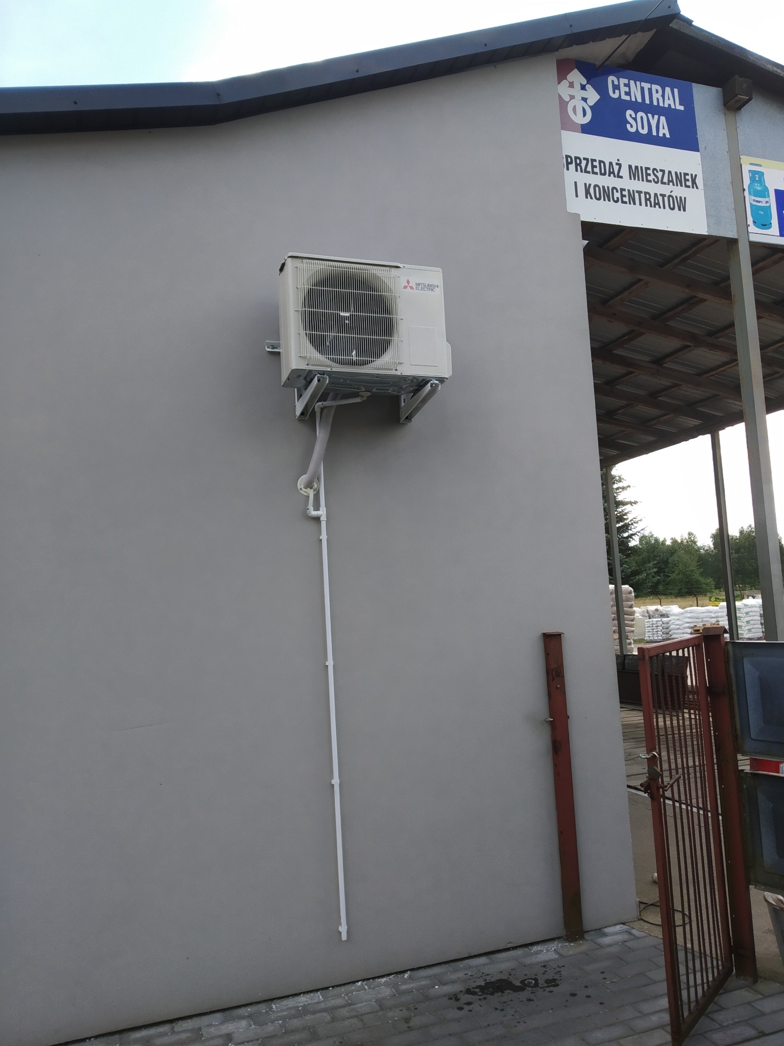 Klimatyzator Mitsubishi Electric zamontowany na szarej ścianie budynku gospodarczego z widoczną instalacją odprowadzającą skropliny, na tle magazynu Central Soya.