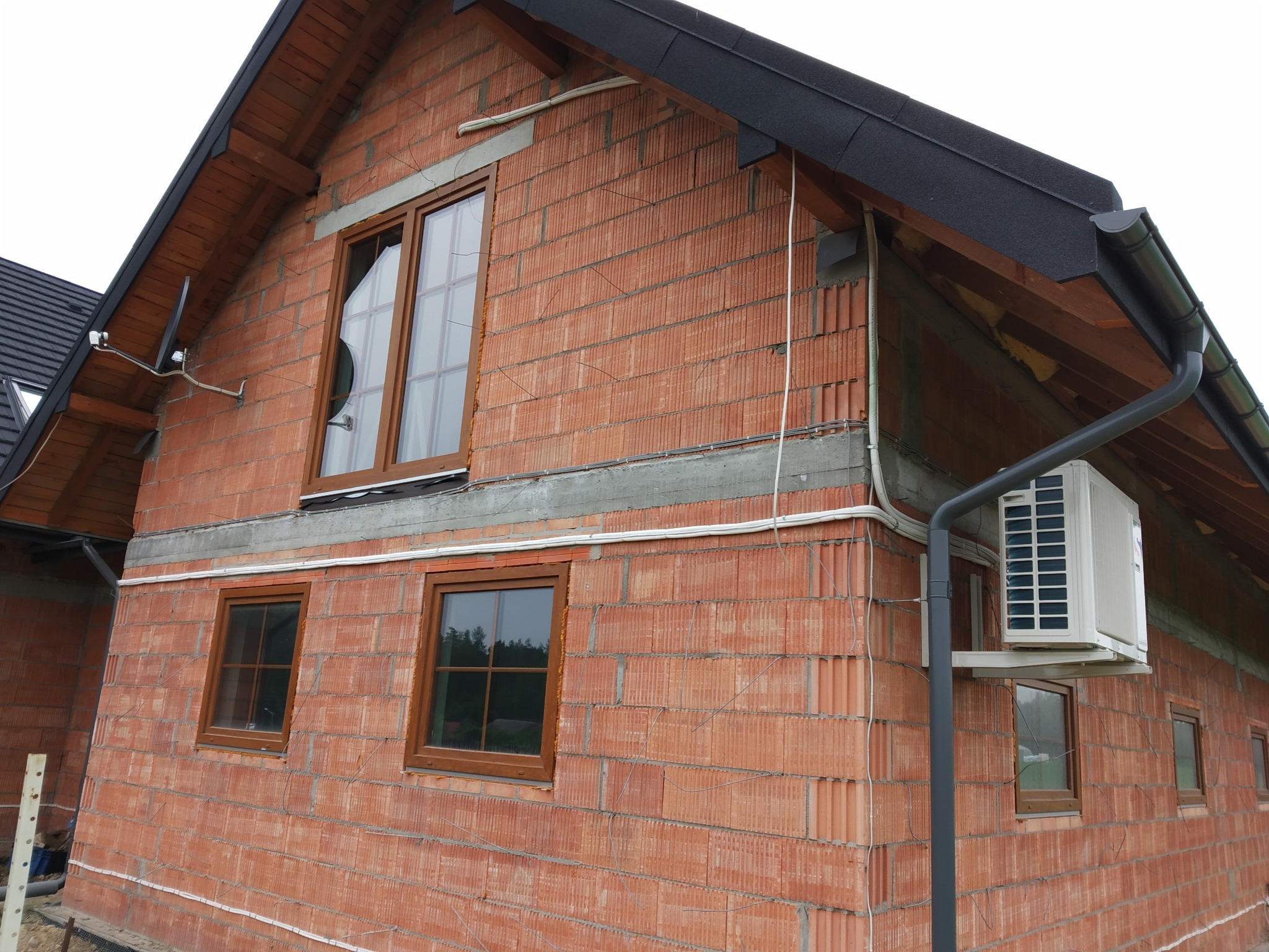 Niedokończona elewacja budynku z czerwonej cegły z zamontowanym zewnętrznym modułem klimatyzacji, widoczne okna z brązowymi ramami, orynnowanie i przewody elektryczne.