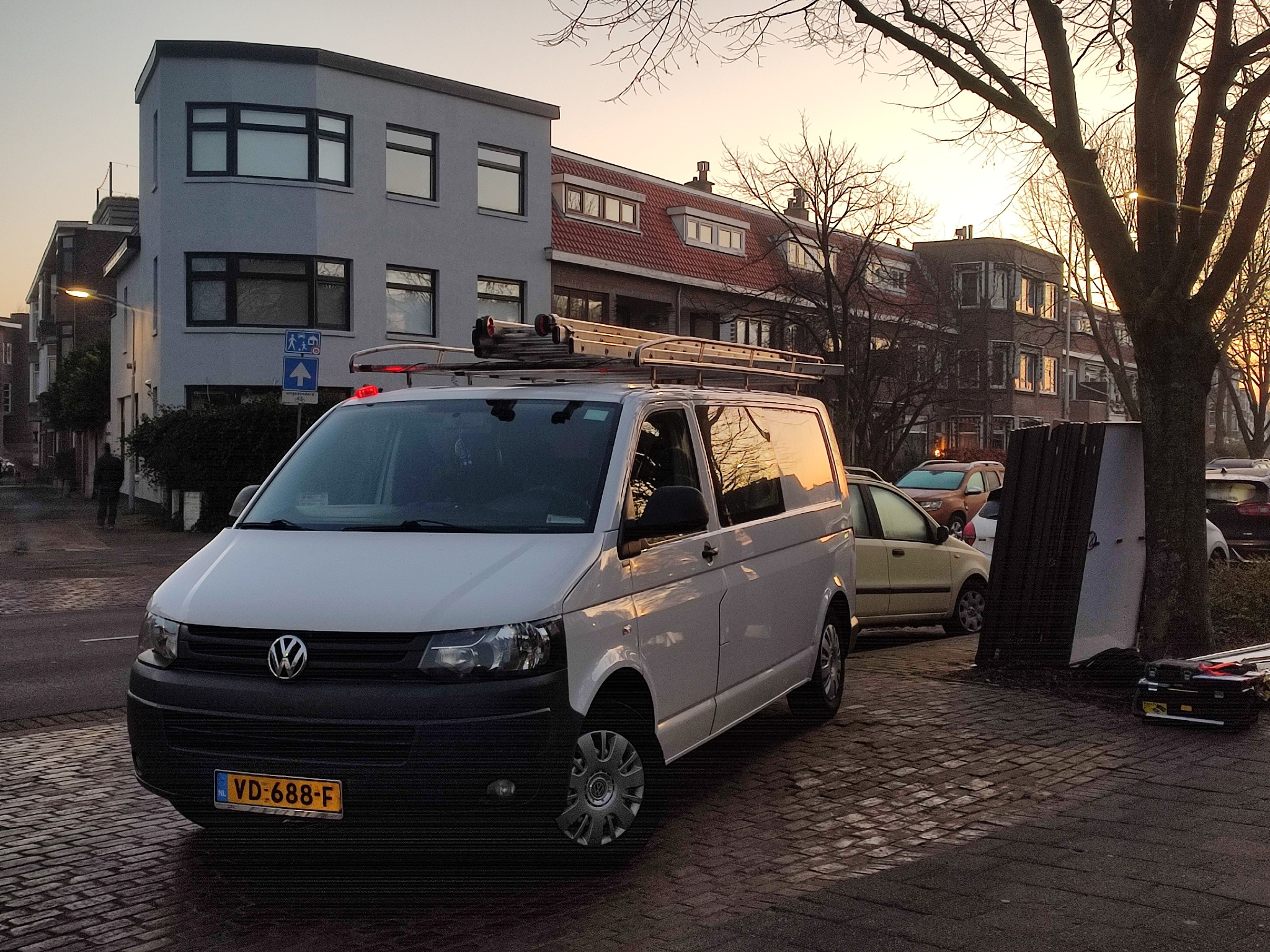 Biały van Volkswagen z drabiną na dachu zaparkowany na brukowanej ulicy w dzielnicy mieszkalnej o zmierzchu, widoczna tablica rejestracyjna VD-688-F.