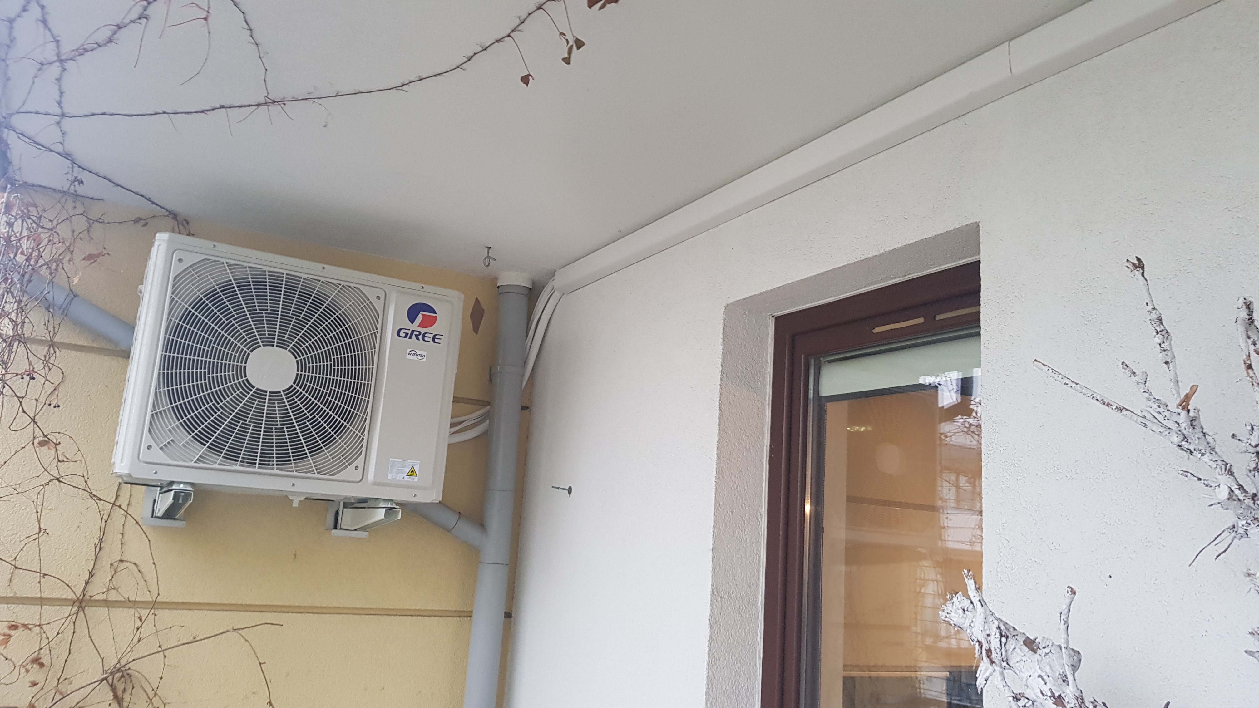 Zewnętrzna jednostka klimatyzacji marki Gree zamontowana na żółtej elewacji domu, widoczne rury odprowadzające skropliny i fragment okna z brązową ramą.