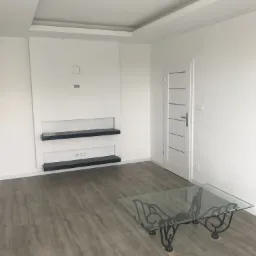 Jasne wnętrze pokoju po remoncie z panelami podłogowymi, białymi ścianami i wnęką z czarnymi półkami oraz białą, nowoczesną stolarką drzwiową i szklanym stolikiem kawowym.