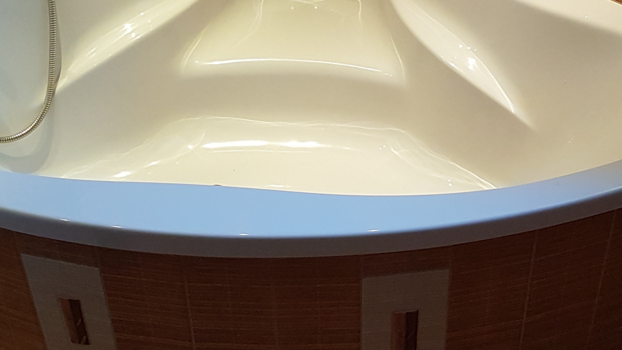 Narożna wanna akrylowa z obudową wykończoną brązowymi płytkami z dekoracyjnymi wstawkami, widoczny chromowany wąż prysznicowy.