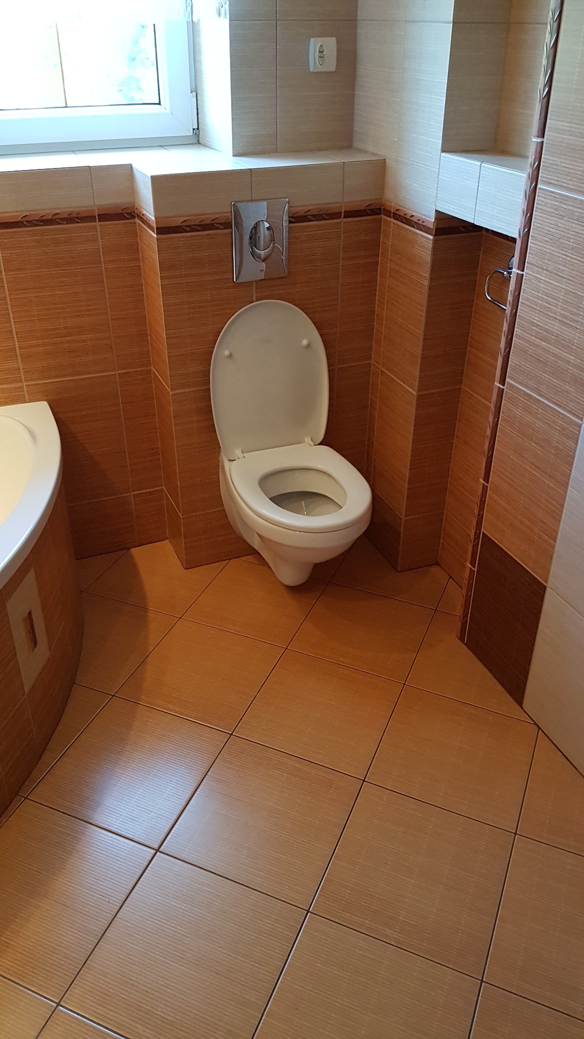 Toaleta z otwartą klapą w łazience wyłożonej brązowymi płytkami, widoczna bateria podtynkowa i fragment wanny.