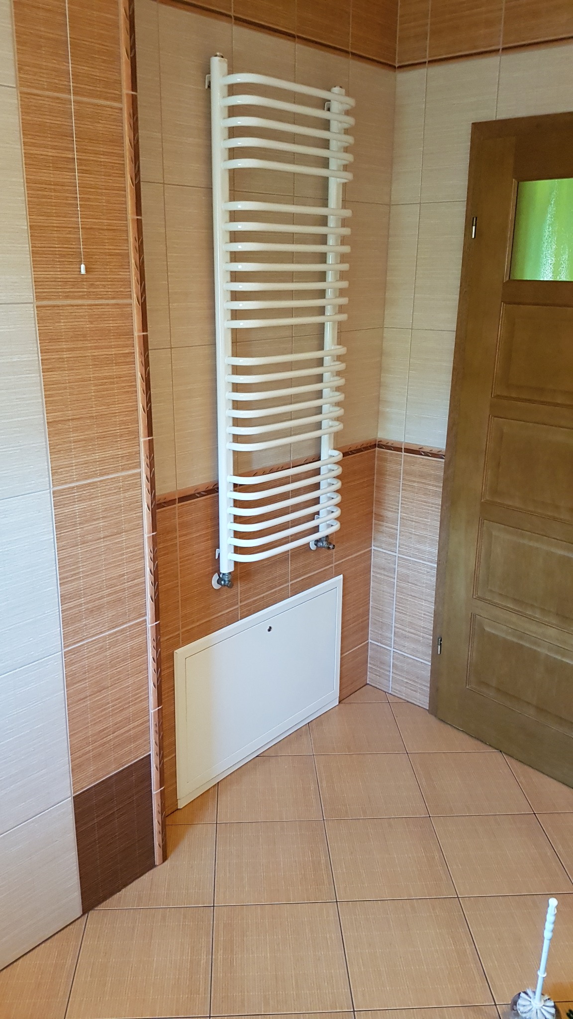 Biały grzejnik drabinkowy zamontowany na brązowo-białej ścianie w łazience, z białą klapą rewizyjną pod nim i drewnianymi drzwiami z przeszkleniem w tle.