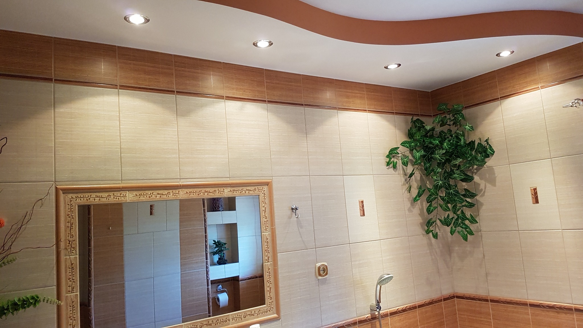 Wykończona łazienka z beżowymi i brązowymi płytkami, lustrem w zdobionej ramie, dekoracyjną rośliną i punktowym oświetleniem sufitowym.