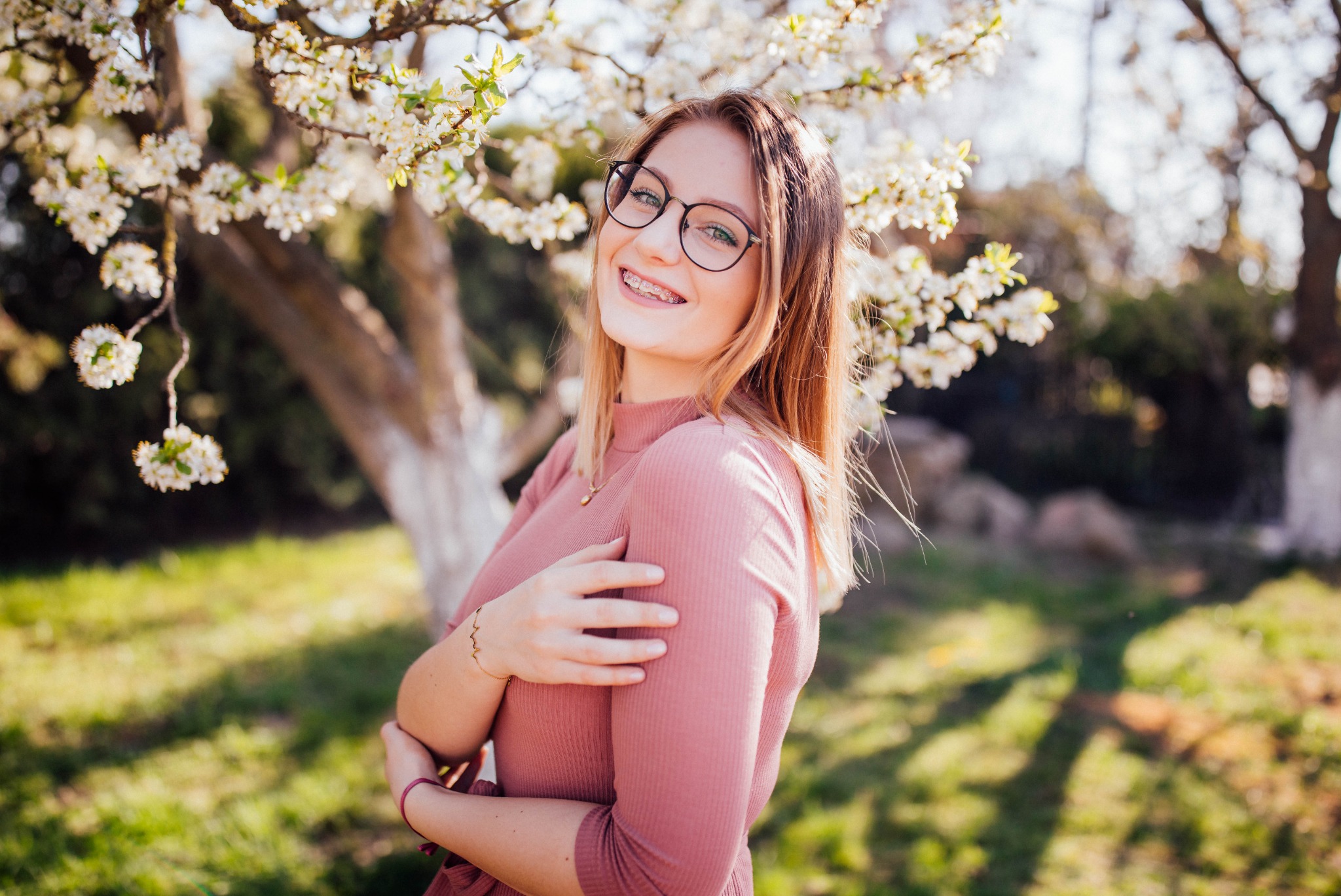 Uśmiechnięta młoda kobieta w okularach i aparacie ortodontycznym, pozująca na tle kwitnącego drzewa owocowego w słoneczny dzień, z rękami skrzyżowanymi na piersiach i złotą bransoletką na nadgarstku.