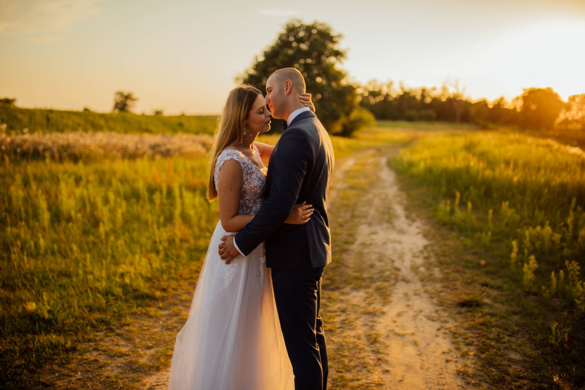 Para młoda obejmująca się na polnej drodze o zachodzie słońca, panna młoda w koronkowej sukni, pan młody w ciemnym garniturze.