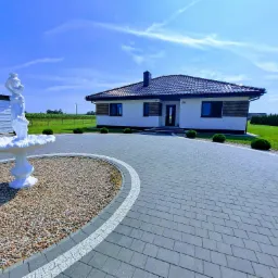 Nowoczesny dom jednorodzinny z szarym brukowanym podjazdem, fontanną z białą figurą kobiety i zadbanym trawnikiem z krzewami.