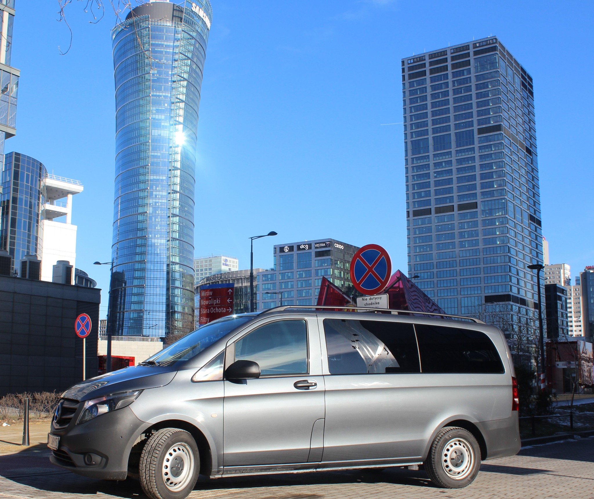 Szary van marki Mercedes-Benz V-Class na tle nowoczesnej architektury Warszawy, w tle widoczne wieżowce i znaki drogowe.