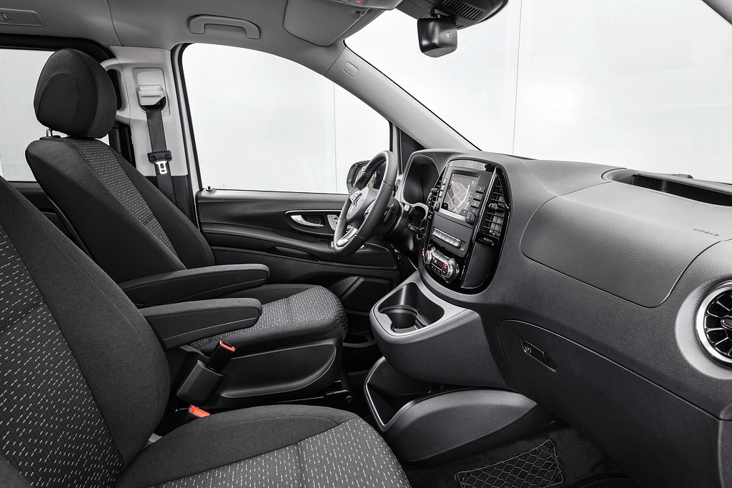 Wnętrze samochodu dostawczego z szarą tapicerką, widoczne fotele z podłokietnikami, deska rozdzielcza z nawigacją i elementami sterowania