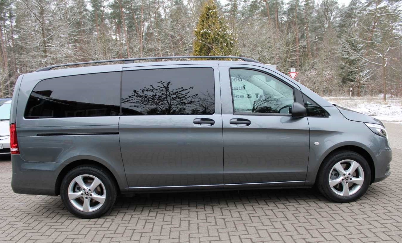 Szary van z przyciemnianymi szybami zaparkowany na brukowanej nawierzchni, z widocznym odbiciem drzew w oknach.