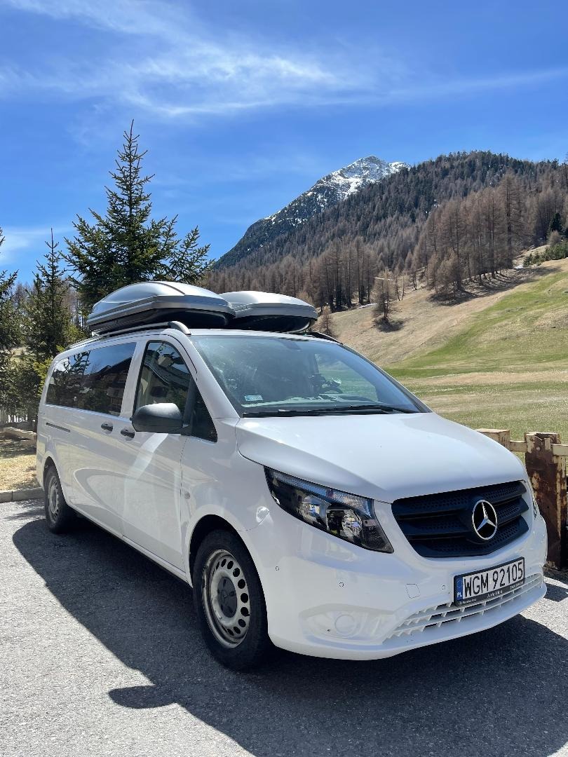 Biały van Mercedes z dwoma bagażnikami dachowymi na tle górskiego krajobrazu z ośnieżonym szczytem.