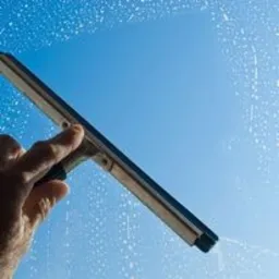 Dłoń z gumową ściągaczką czyści szklaną powierzchnię, pozostawiając smugi i krople wody na tle błękitnego nieba.