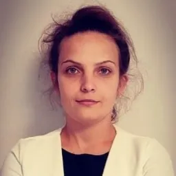 Portret kobiety w białej marynarce i czarnej bluzce na jasnym tle, z włosami upiętymi do góry.