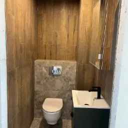 Nowoczesna łazienka z drewnopodobnymi płytkami ściennymi, marmurową mozaiką za toaletą, wiszącą szafką z umywalką w kolorze butelkowej zieleni i czarną baterią.