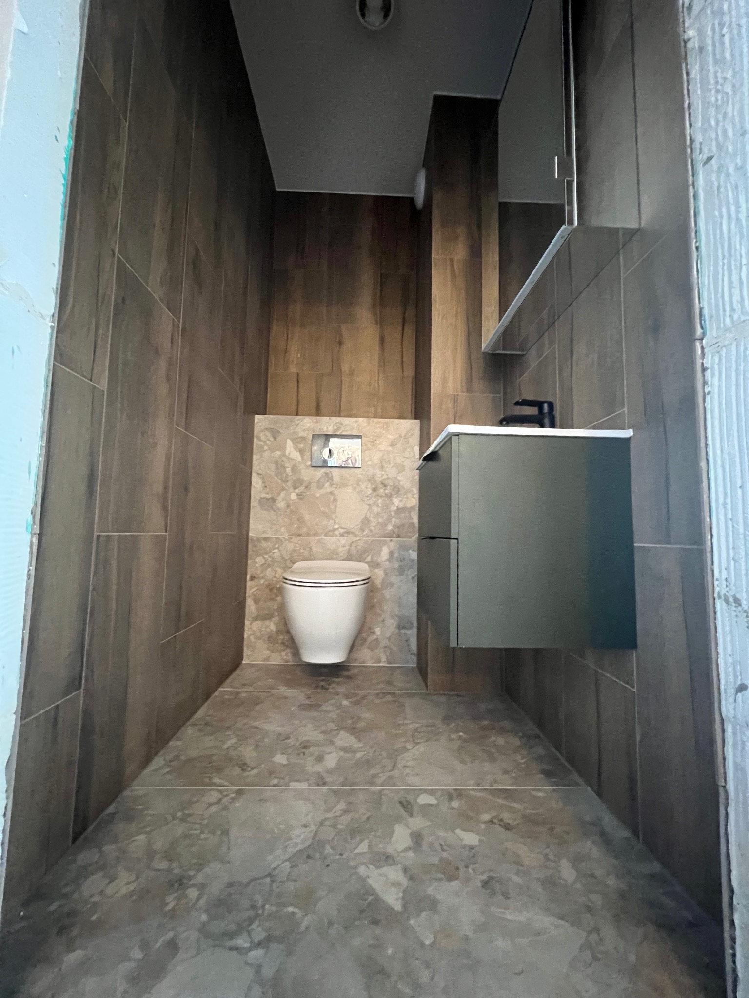 Nowoczesna łazienka w trakcie wykańczania: brązowe płytki imitujące drewno na ścianach, marmurowa podłoga, podwieszana toaleta z przyciskiem spłukującym, wisząca szafka z umywalką i czarną baterią.