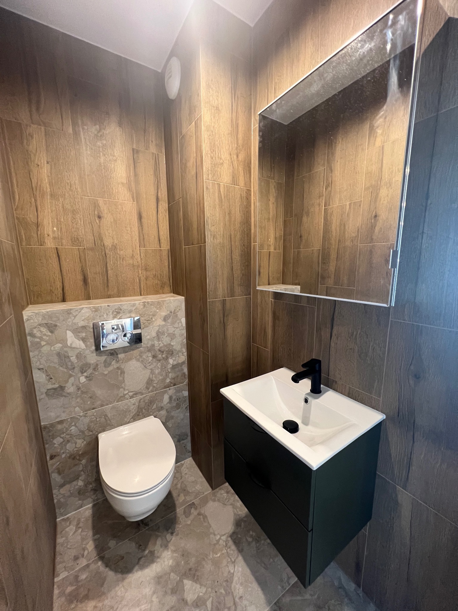 Nowoczesna łazienka z drewnopodobnymi płytkami ściennymi, szarą mozaiką za toaletą, białą miską ustępową, umywalką nablatową z czarną armaturą i szafką w kolorze butelkowej zieleni oraz lustrem.