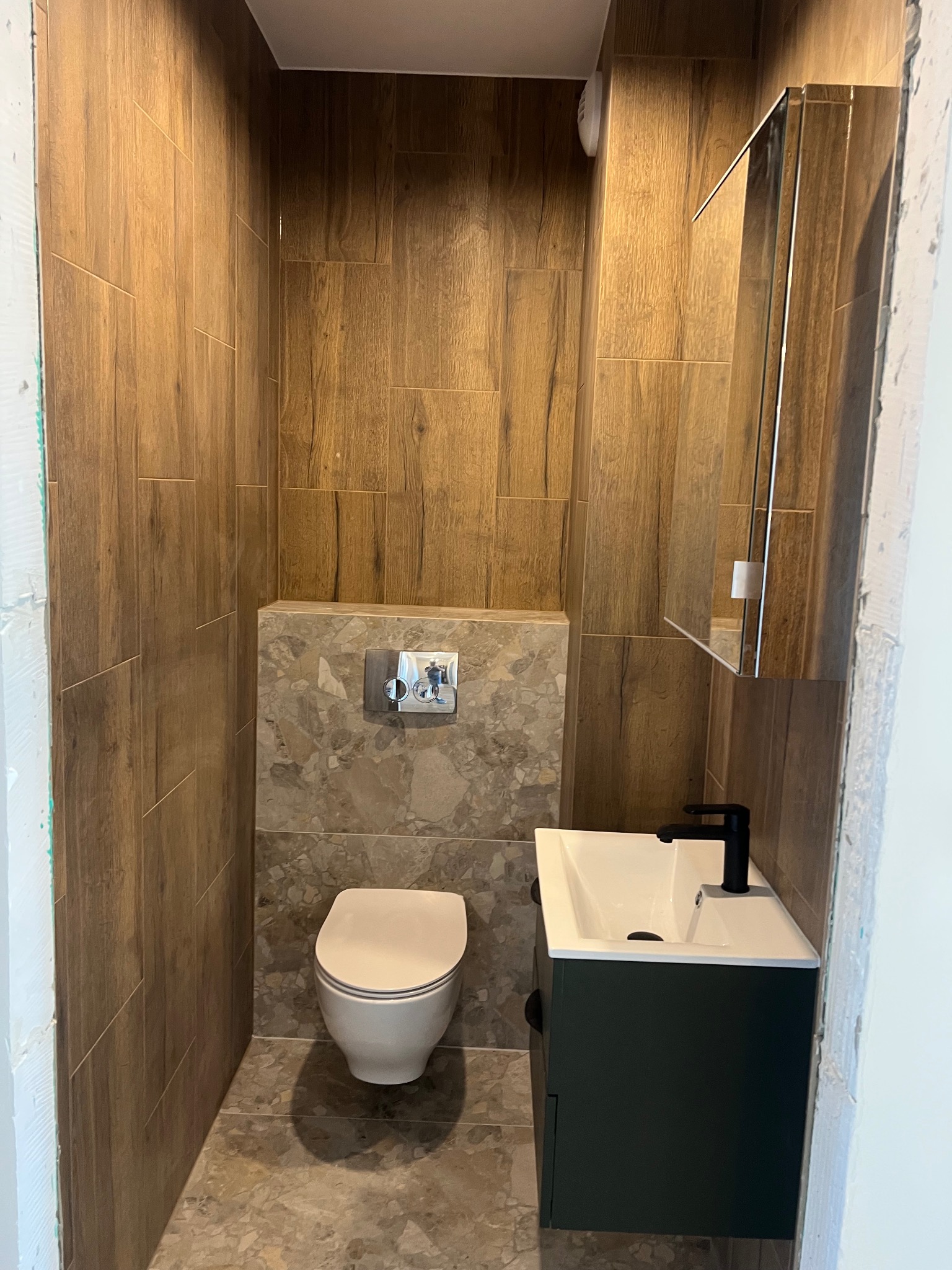 Nowoczesna łazienka z drewnopodobnymi płytkami ściennymi, marmurową mozaiką za toaletą, wiszącą szafką z umywalką w kolorze butelkowej zieleni i czarną baterią.