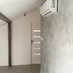 Biały klimatyzator ścienny zamontowany w pokoju z szarą, dekoracyjną ścianą, drewnianą podłogą i drzwiami z przeszkleniami.