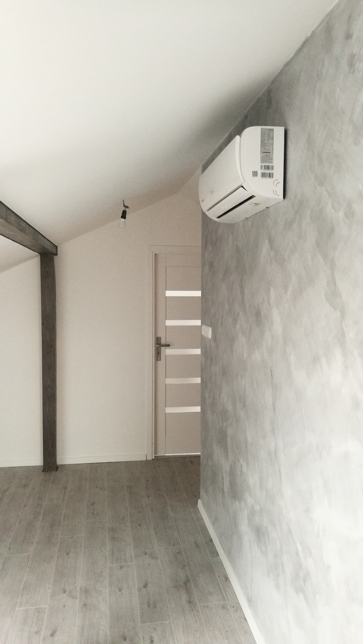 Biały klimatyzator ścienny zamontowany w pokoju z szarą, dekoracyjną ścianą, drewnianą podłogą i drzwiami z przeszkleniami.