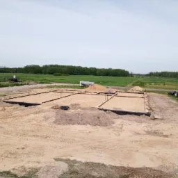 Przygotowany teren pod fundamenty domu jednorodzinnego, widoczne szalunki i zbrojenie, w tle krajobraz wiejski.