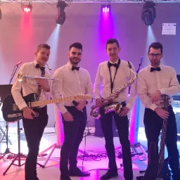 Czteroosobowy zespół muzyczny w białych koszulach i muchach, z instrumentami: gitara, trąbka, saksofon i gitara basowa, na scenie z oświetleniem w kolorach czerwonym i niebieskim.