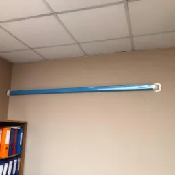 Błękitna rura wentylacyjna zamontowana przy suficie w pomieszczeniu biurowym, widoczny fragment regału z segregatorami.