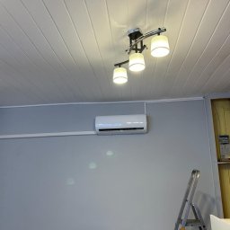 ULTECH Jakub Ulman - Biały klimatyzator zamontowany na szarej ścianie w pomieszczeniu z podwieszanym sufitem, widoczna drabina i częściowo otwarta szafka z instalacją elektryczną.