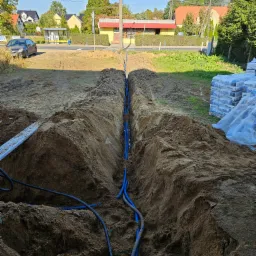 Wykop w ziemi z ułożonymi niebieskimi rurami karbowanymi, widoczne worki z zaprawą budowlaną w tle, domy jednorodzinne i słup elektryczny w oddali.
