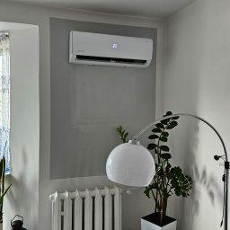 ULTECH Jakub Ulman - Biały klimatyzator ścienny z wyświetlaczem temperatury, zamontowany na szarej ścianie w salonie w Górze, widoczny obok grzejnika i lampy podłogowej, z rośliną doniczkową w tle.