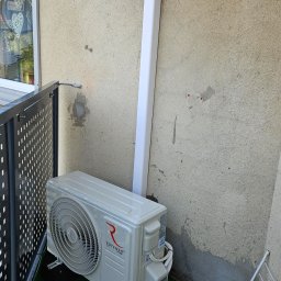 ULTECH Jakub Ulman - Zewnętrzna jednostka klimatyzacji Rotenso Ukura zamontowana na balkonie z imitacją trawy, widoczna biała rura odprowadzająca skropliny oraz ślady po wcześniejszych mocowaniach na ścianie.