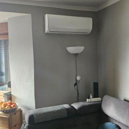 ULTECH Jakub Ulman - Biała jednostka klimatyzacyjna zamontowana na szarej ścianie w pokoju, widoczna nad szarą sofą z poduszką i lampą stojącą, z owocami w misce w tle.