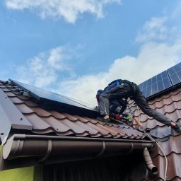 ULTECH Jakub Ulman - Montaż paneli słonecznych na brązowym dachu z dachówki ceramicznej, widoczna antena telewizyjna i fragment zielonej ściany budynku.