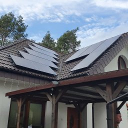 ULTECH Jakub Ulman - Panele fotowoltaiczne na brązowym dachu domu jednorodzinnego, widok z dołu na tle błękitnego nieba z chmurami, zadaszony taras i osoba w tle.