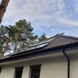 Panele fotowoltaiczne zamontowane na dachu domu jednorodzinnego z brązową dachówką, widoczne z perspektywy ogrodu z drzewami i pnączami.