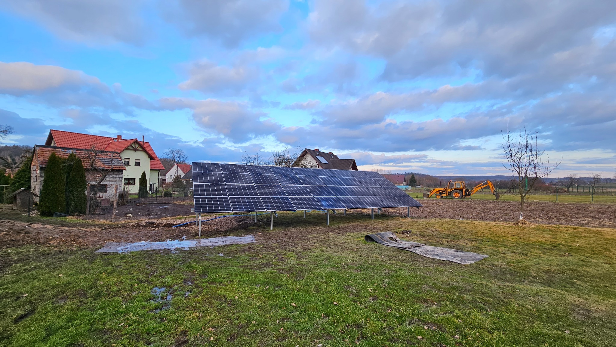 Panel fotowoltaiczny na metalowych podporach, w tle domy, traktor i pochmurne niebo o zmierzchu.