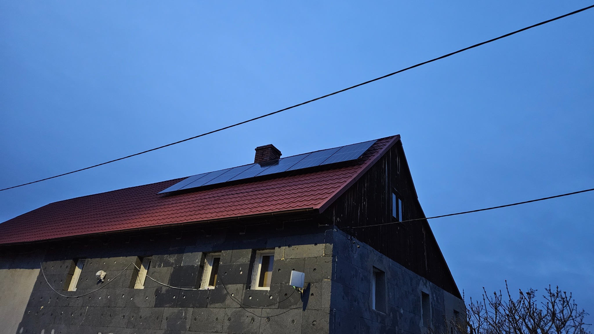 Dom z czerwoną dachówką i panelami fotowoltaicznymi na dachu, w trakcie ocieplania styropianem, widoczne okna i przewody elektryczne na tle błękitnego nieba.